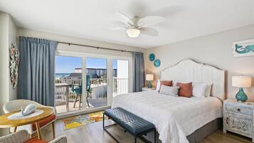 A hotel room with a bed, a bench, a small table, and a chair. There is a balcony with a view of the beach.