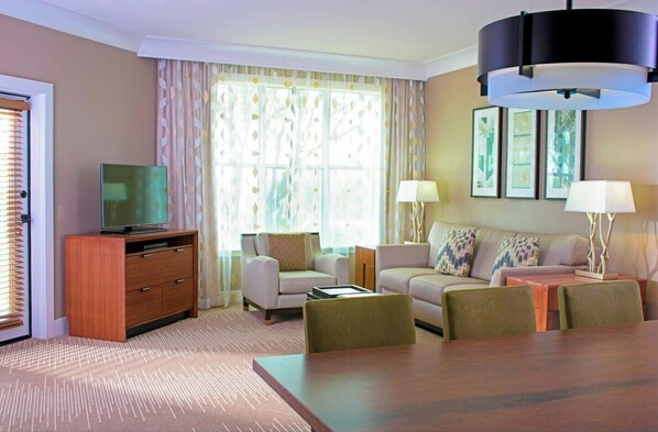 Ein Hotelzimmer mit Fernseher, einer Couch, einem Esstisch und einem großen Fenster mit Vorhängen.