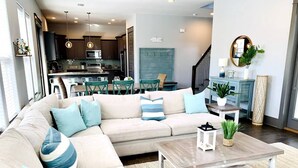 A modern living room with a beige sectional sofa, a wooden coffee table, and a kitchen in the background.
