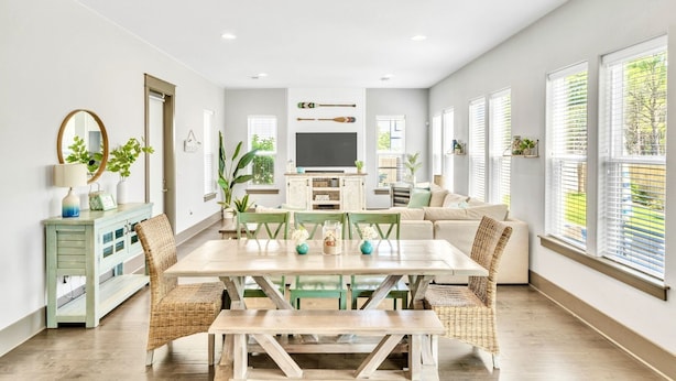 A spacious living area with a dining table, wicker chairs, a sofa, and large windows with blinds.