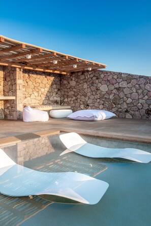 A stone wall with a wooden pergola and a pool area with white lounge chairs and pillows. - Villa Retreat (Milos)