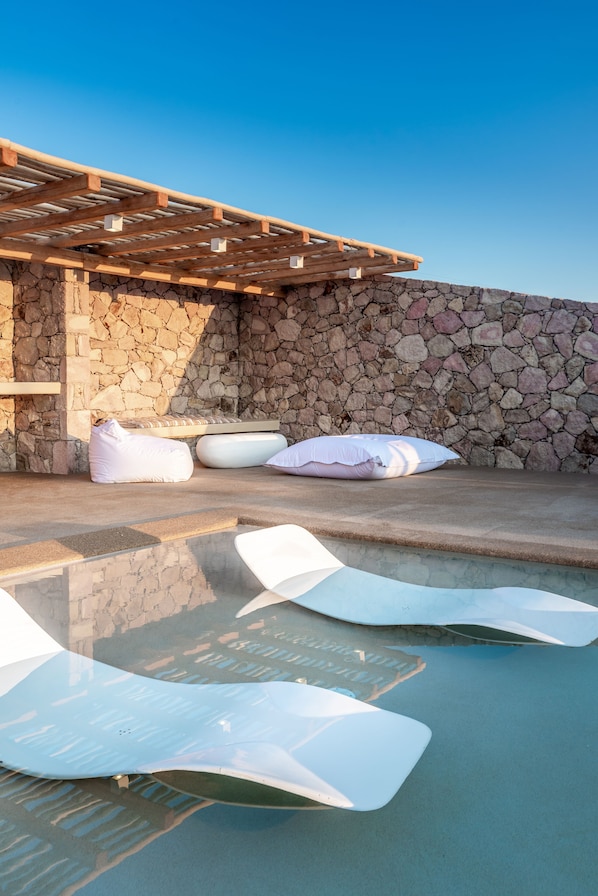 Un muro de piedra con una pérgola de madera y una zona de piscina con sillones y cojines blancos.