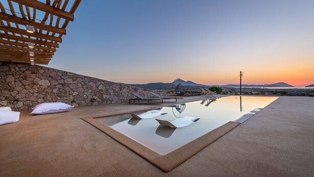 An infinity pool with a stone wall and lounge chairs, set against a sunset view. - Villa Retreat (Milos)