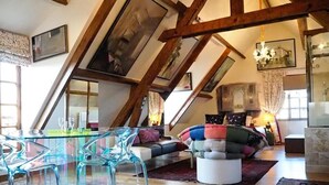 Un loft spacieux avec de hauts plafonds, des poutres apparentes, une table à manger en verre et un coin salon confortable agrémenté de coussins colorés.