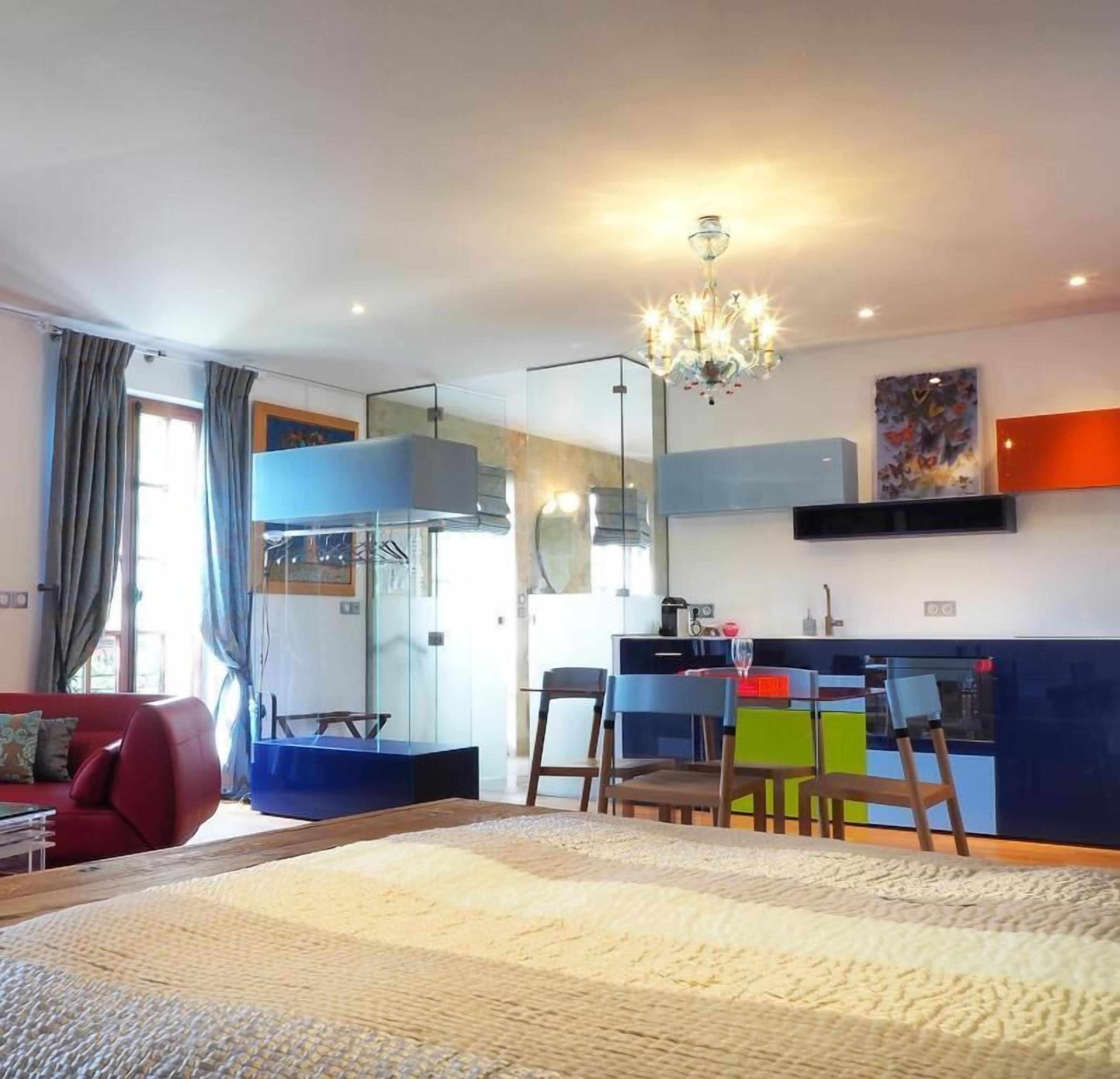 Ein modernes Wohnzimmer mit einem großen Bett, einem roten Sofa, einem Essbereich mit bunten Stühlen und einer Küche im Hintergrund.