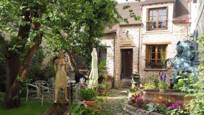 Une cour avec un chemin en pierre, des plantes en pot, une statue et une maison rustique aux murs de pierre et aux volets en bois.