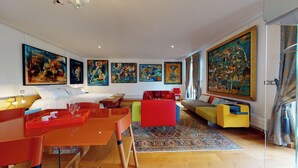 Une chambre d’hôtel moderne avec un grand lit, un coin repas avec une table rouge, des canapés colorés et plusieurs tableaux abstraits accrochés aux murs.