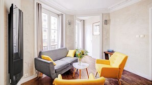 Een woonkamer met een grijze bank, gele fauteuils, een kleine ronde tafel met een bloevas, en een televisie links.