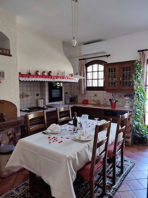 Un coin repas avec une table dressée pour quatre personnes, un four en briques et une cuisine dotée de meubles en bois et d’un plan de travail carrelé.