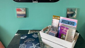 Una pequeña mesa con un libro, crucigrama y una revista, con una ventana que muestra una vista de una masa de agua y barcos.