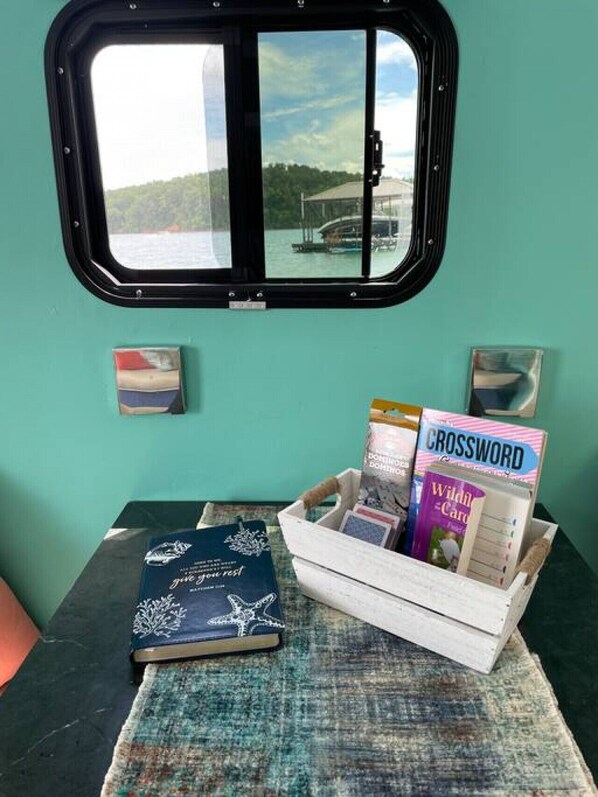 A small table with a book, crossword puzzle, and a magazine, with a window above displaying a view of a body of water and boats.