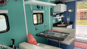 Un interior compacto y bien iluminado, con una pared color turquesa, una pequeña kitchenette, una zona de comedor con mesa y banco, y un ventanal que deja entrar luz natural.