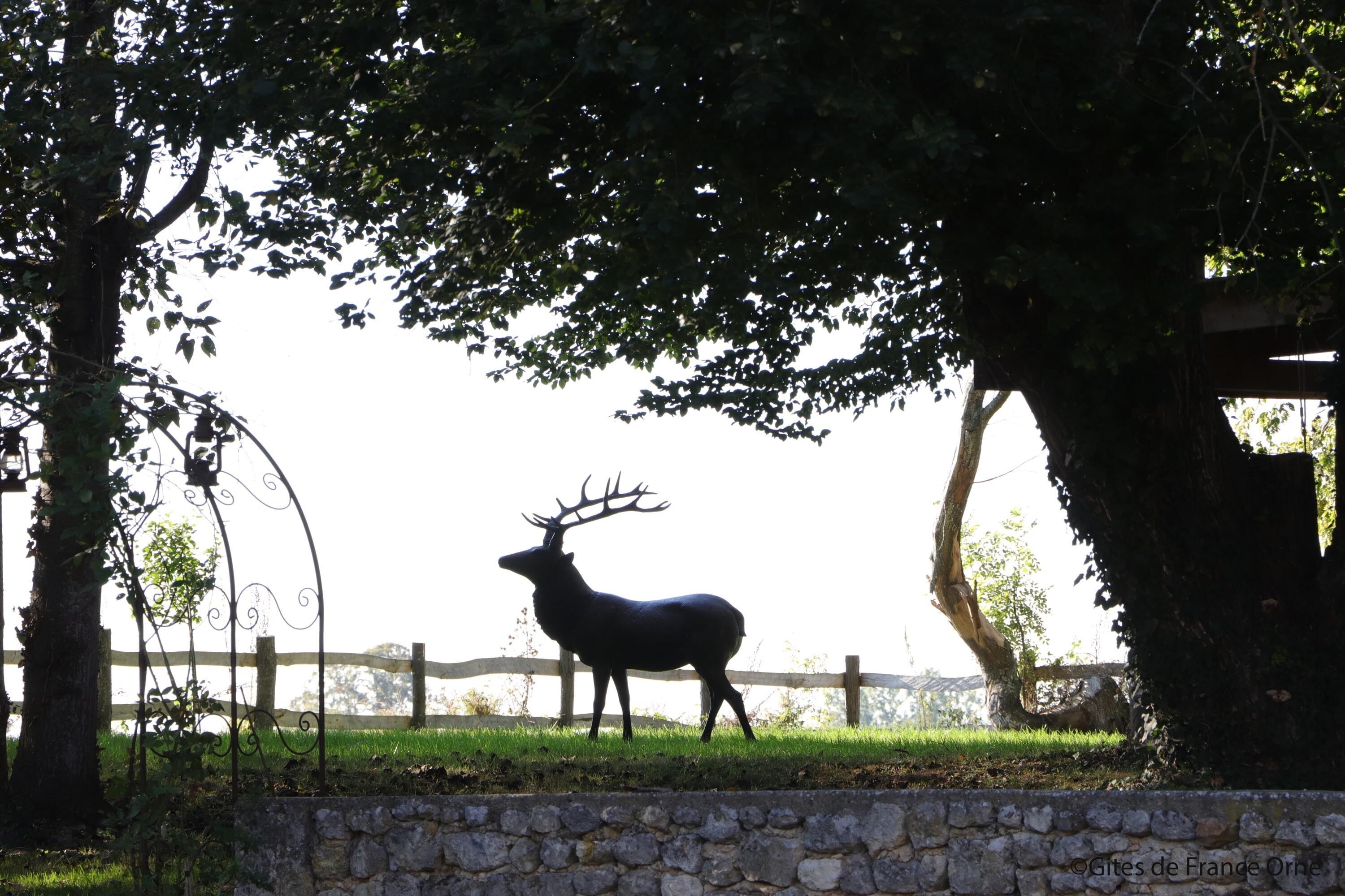 A deer statue in a park with a stone wall and wooden fence.