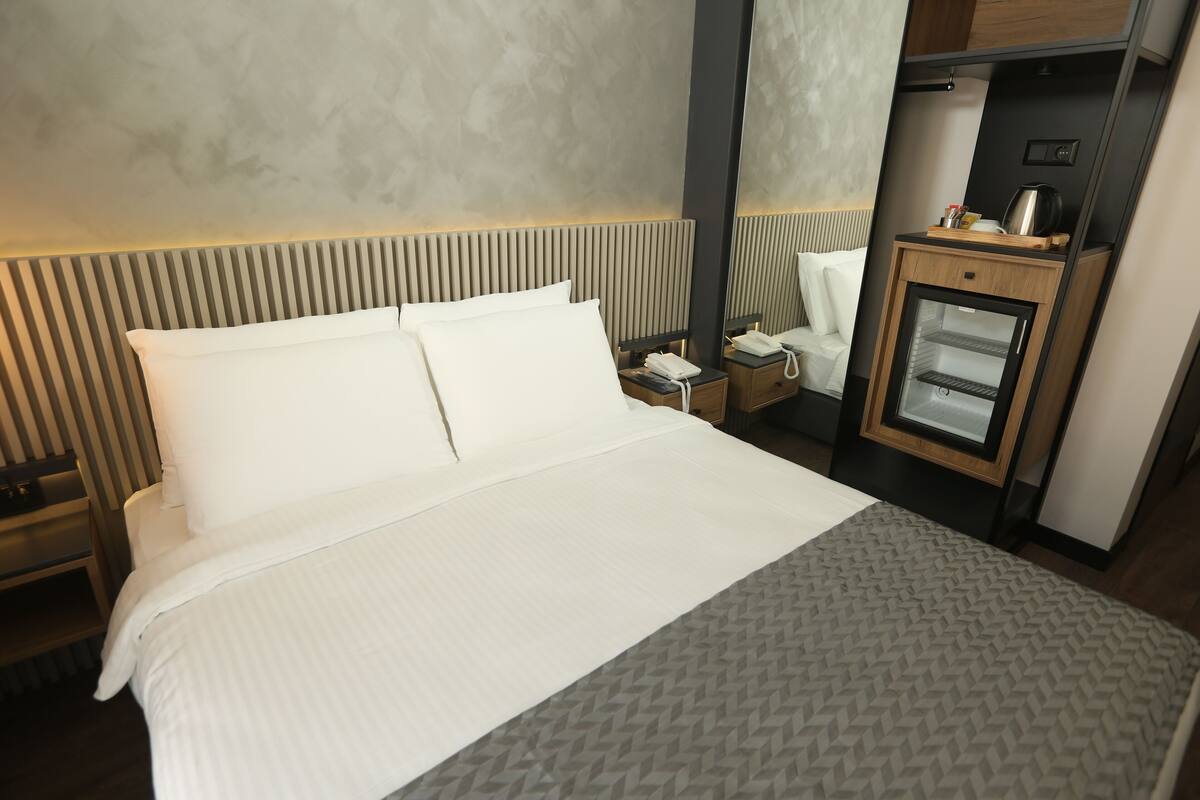 A hotel room with a large bed, a bedside table with a lamp, a small refrigerator, and a mirror.