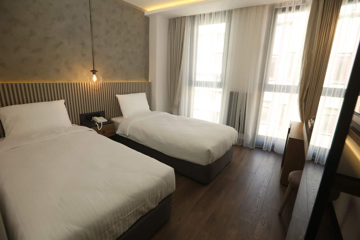 A hotel room with two beds, a nightstand, a lamp, and a window with curtains.