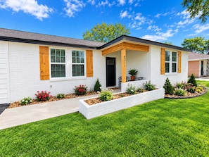 A modern single-story house with a white exterior, a front porch with wooden shutters, and a well-maintained front yard with various plants and a small tree.