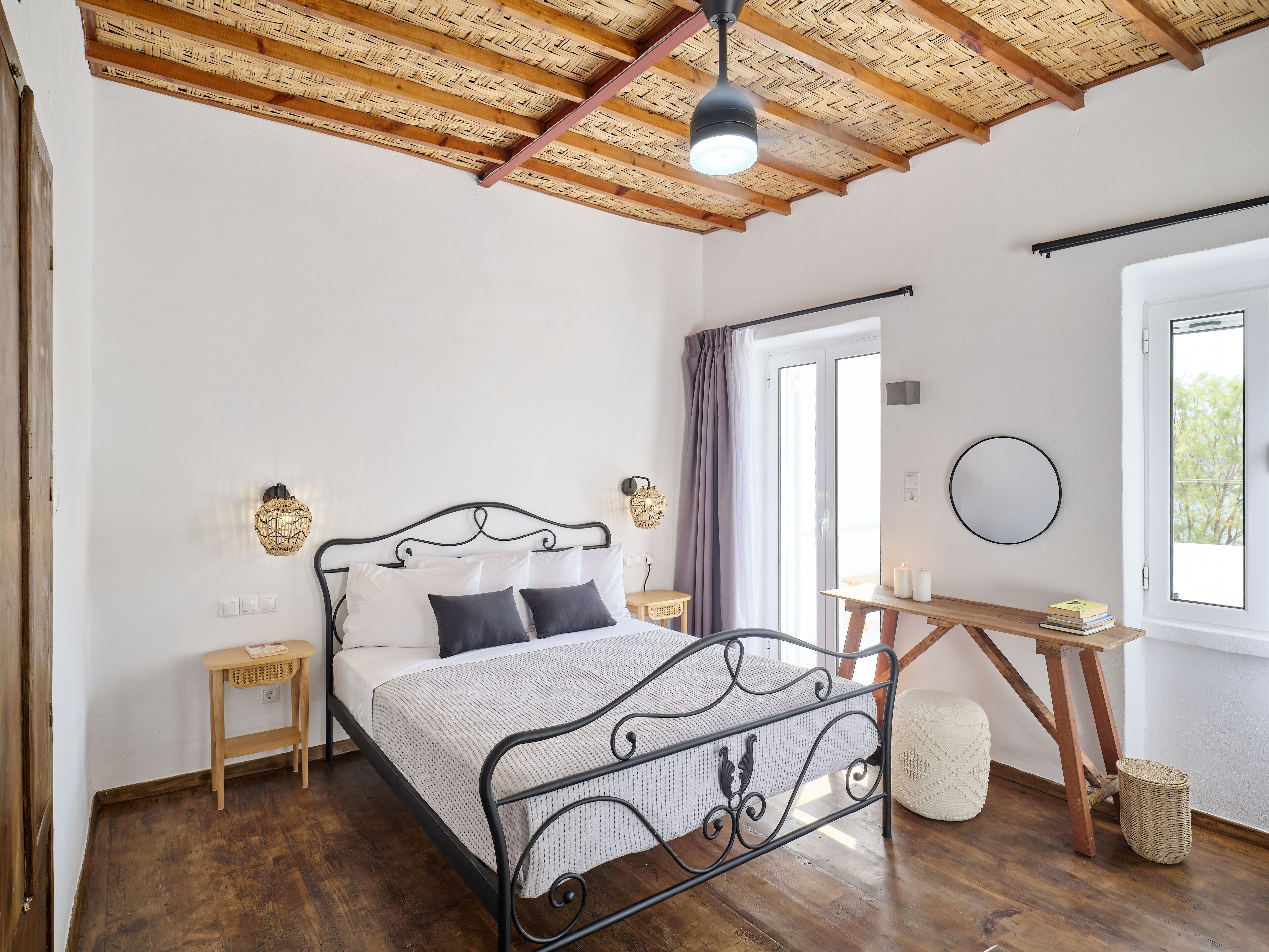 Un dormitorio con cama, mesitas de noche, escritorio, silla, ventana y techo con vigas de madera.