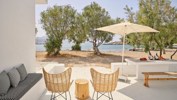 Een terras met twee rieten stoelen, een houten tafel en een bankje. Een witte muur en uitzicht op zee en bomen.
