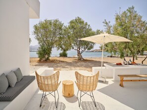 Une terrasse avec deux chaises en osier, une table en bois et un banc. On y trouve un mur blanc et une vue sur la mer et les arbres.