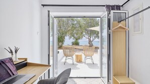 Een moderne kamer met een laptop op een bureau, een grijze bank en uitzicht op een strand met tuinmeubilair.
