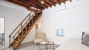 Een kamer met een houten trap, een bed, een stoel, een klein tafeltje en vloertegels met een patroon.