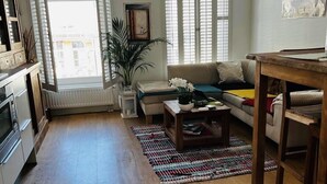 A spacious living room with a sofa, coffee table, and a dining table. There is a large window with blinds, a radiator, and a potted plant. - Private rooftop 35m2, high-end apartment (Amsterdam)