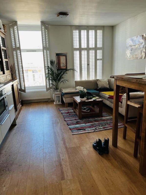 Uma sala de estar ampla com sofá, mesa de centro e mesa de jantar. Há uma janela grande com persianas, um radiador e um vaso de planta.