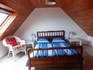 Ein Schlafzimmer mit schrägen Wänden, einem hölzernen Bett mit blauer Bettwäsche, einem weißen Stuhl mit rotem Kissen und zwei Nachttischlampen.