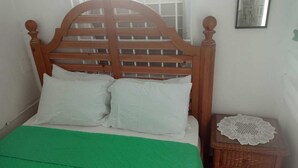 Una cama con cabecera de madera, dos almohadas blancas y una colcha verde. Hay una mesita de noche de madera con un mantel blanco encima.