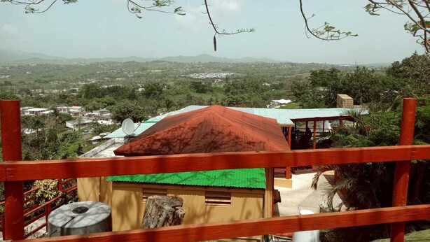 Vista de un paisaje rural con una pequeña construcción de techo rojo y toldo verde, rodeada de abundante vegetación y colinas distantes.