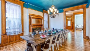 Une salle à manger avec une table en bois dressée pour six personnes, des chaises blanches, un lustre et une cheminée.