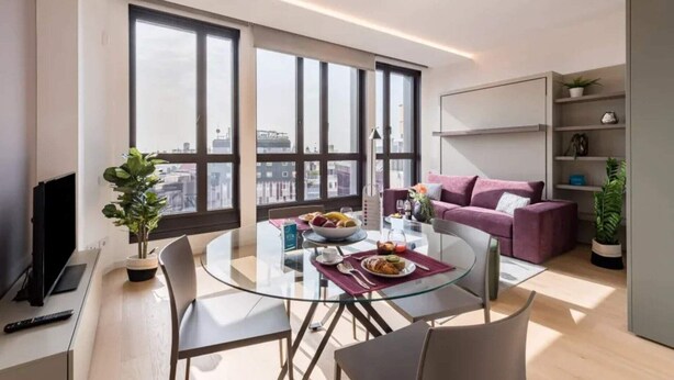 A modern living room with a glass dining table, a purple sofa, and large windows offering a city view. - Sweett | Central tower I (Milano)