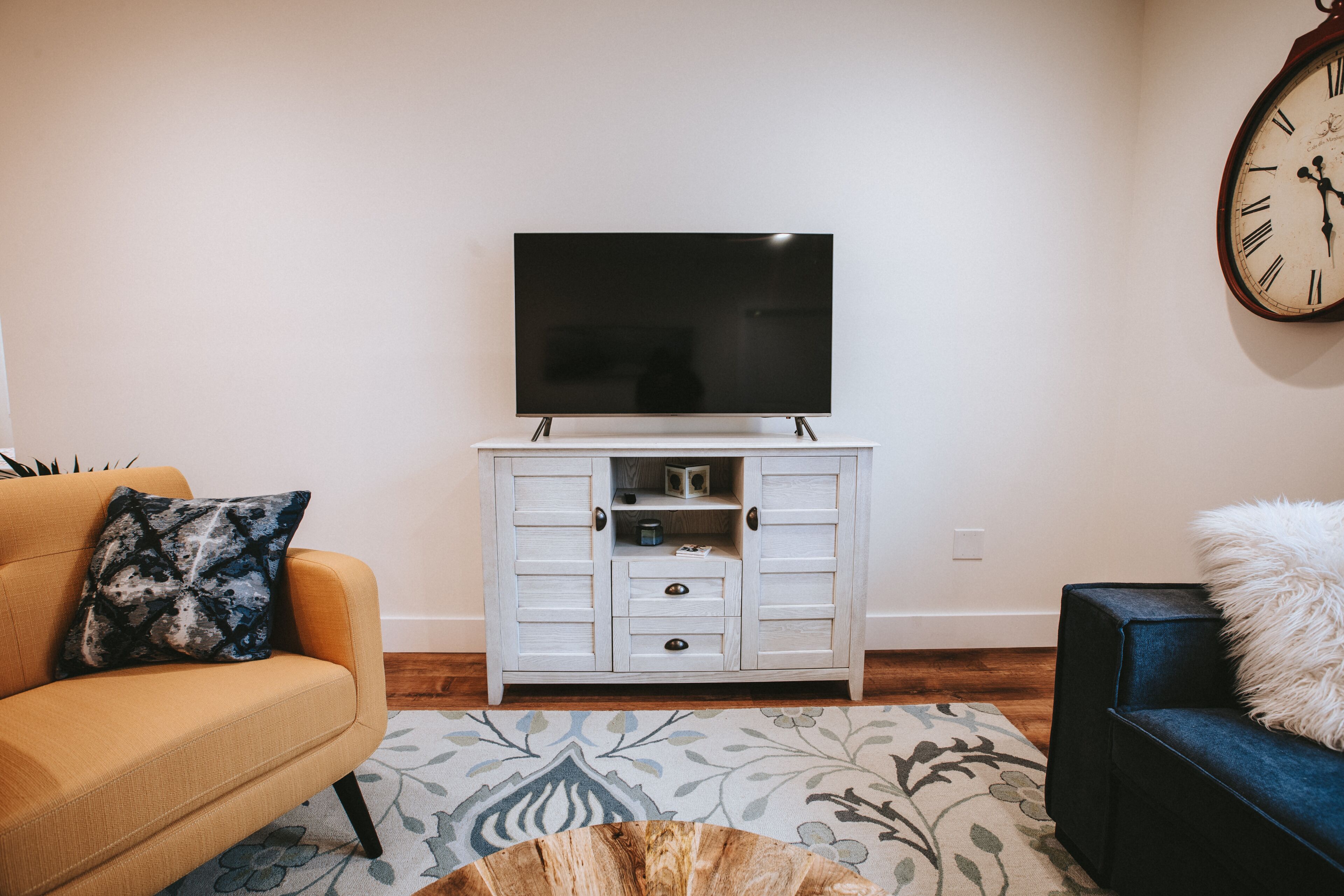 Una sala de estar con un televisor de pantalla plana sobre un mueble blanco, un sofá con un cojín estampado y un sillón azul con un cojín mullido.