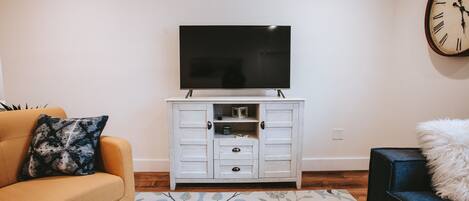 Una sala de estar con un televisor de pantalla plana sobre un mueble blanco, un sofá con un cojín estampado y un sillón azul con un cojín mullido.