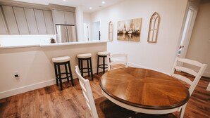 Zona pranzo con tavolo rotondo in legno circondato da sedie bianche, cucina con mobili bianchi e frigorifero in acciaio inossidabile, e un quadro appeso al muro.
