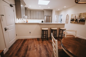 Una cocina con suelo de madera, una puerta blanca y un comedor con mesa de madera y taburetes de bar.