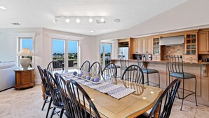 Un comedor amplio con una mesa grande y sillas, una cocina con armarios de madera, y vistas al exterior a través de amplios ventanales.