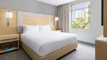 A hotel room with a large bed, a nightstand, a lamp, a window with a view of buildings, and a wall-mounted TV.