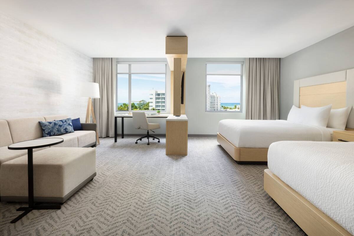 Une chambre d’hôtel équipée d’un canapé, d’un bureau, d’un lit et de grandes fenêtres donnant sur la ville.