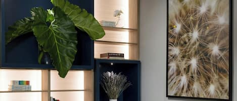 A modern living room with a white sofa, a round coffee table, and a large plant. There is a bookshelf with books and decorative items, and a large framed picture on the wall.