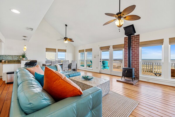 A spacious living room with a blue leather sofa, a coffee table, and a fireplace. There are ceiling fans, large windows, and a view of the outdoors.