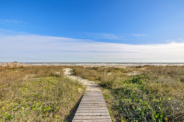 Ein hölzerner Steg führt zu einem Strand mit Dünen und spärlicher Vegetation.
