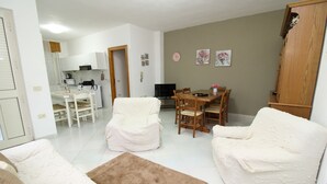 A living room with two white armchairs, a brown rug, a dining table with chairs, and a kitchen in the background. - Janna e Mare Holiday Home (San Giovanni)