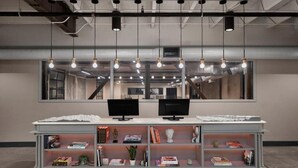 A modern interior with a central desk, two monitors, hanging lights, and a bookshelf. - Evo Hotel (Salt Lake City)