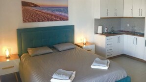 A bedroom with a large bed, a painting of a beach, a kitchenette, and a nightstand with a lamp. - Modern, stylish apt by the sea with large balcony, sea view, close to the center (Sarandë)