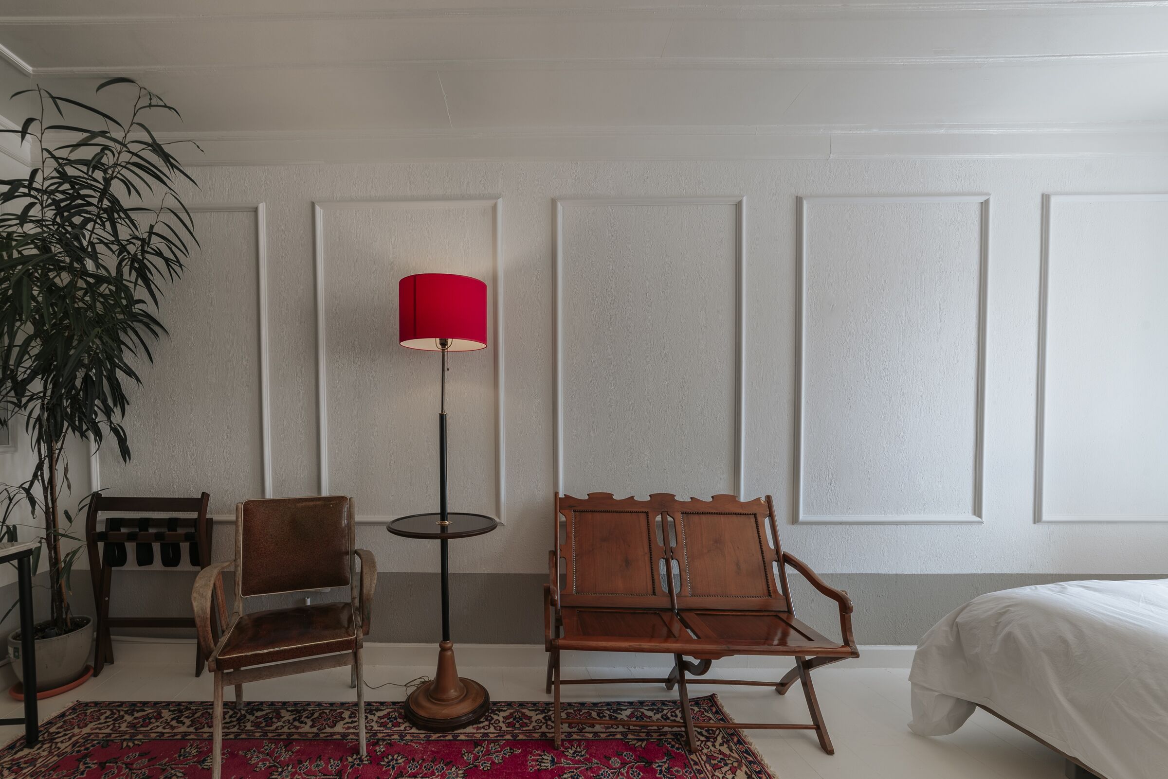 A room with a bed, two wooden chairs, a red floor lamp, a potted plant, and a patterned rug.
