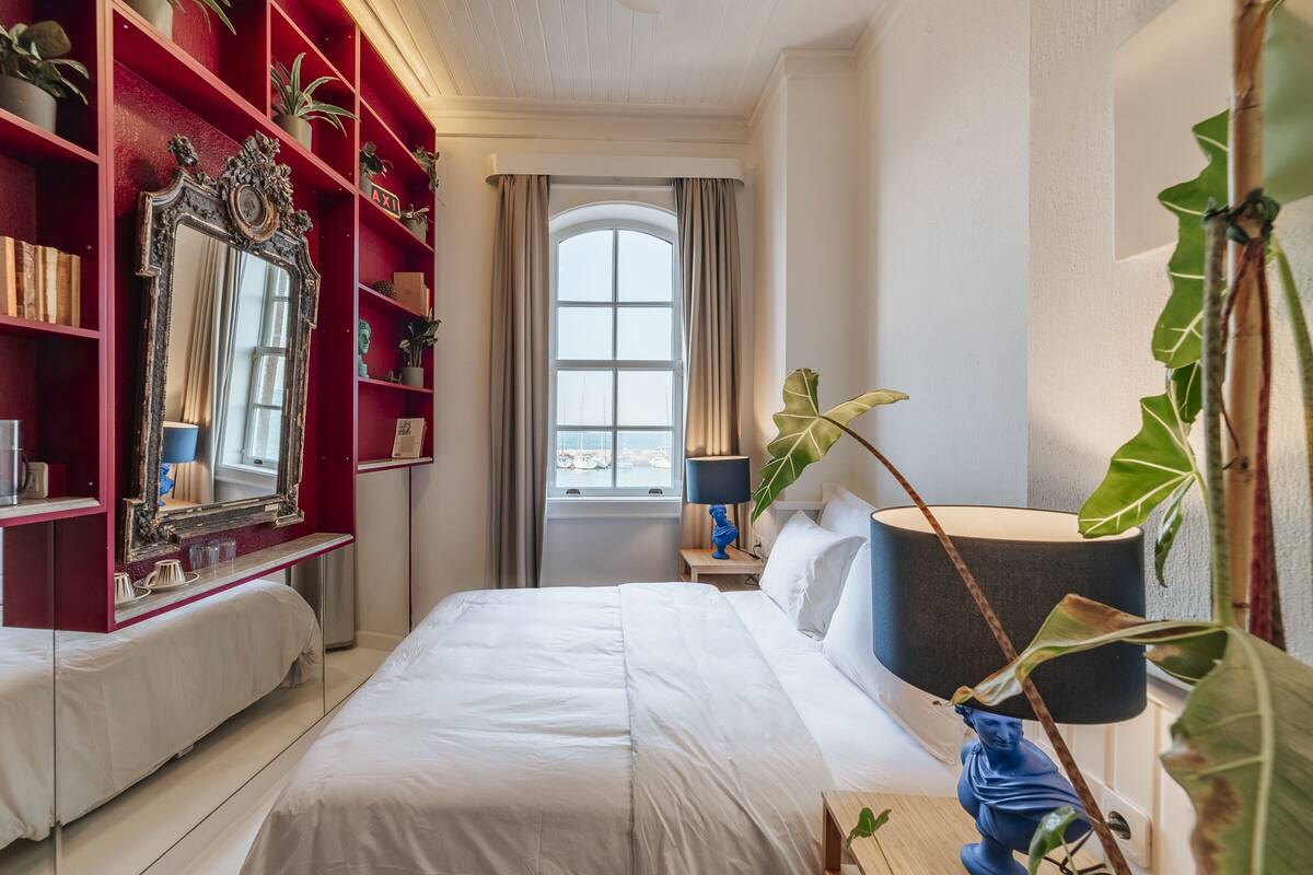 A hotel room with a large bed, a red bookshelf, a mirror, and a window with curtains.