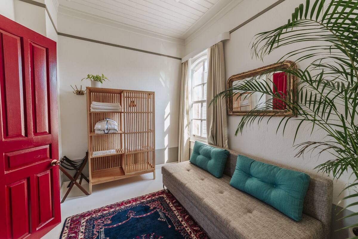 A room with a red door, a wooden wardrobe, a beige sofa with teal cushions, a patterned rug, and a large mirror on the wall.