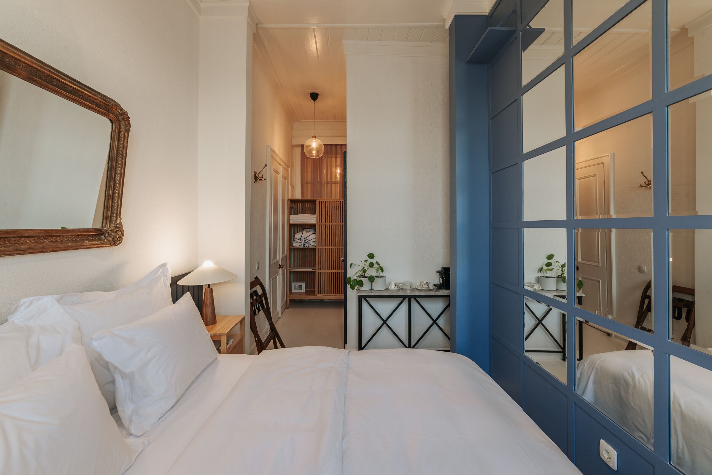 Een slaapkamer met een groot bed, een houten nachtkastje, een spiegel en een uitzicht door een blauw omlijst raam.
