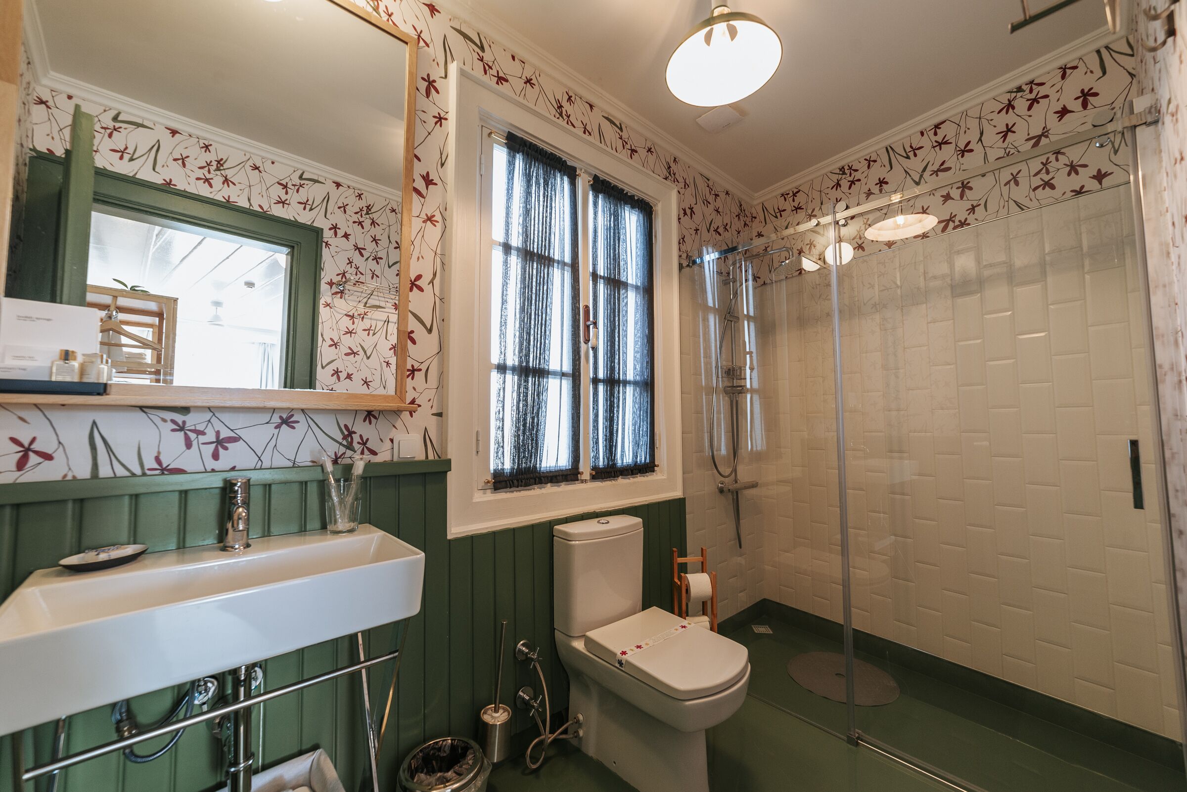 Un baño con lavamanos blanco, inodoro, ducha y ventana con papel tapiz floral.